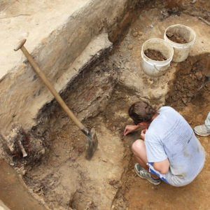 student excavating in the corner of a unit next to a shovel set upright against the unit wall