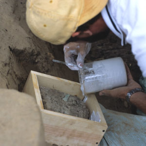 Conservator using a tool to spoon solution from a glass container onto an artifact in a wooden tray