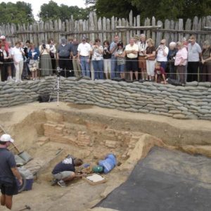 Large group watching two conservators in an excavation unit placing an armor backplate on a tray