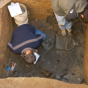 two archaeologists bending over a half-buried human skull in an excavation unit