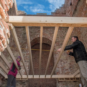 Two workers measure a wooden frame spanning the width of a roofless brick tower
