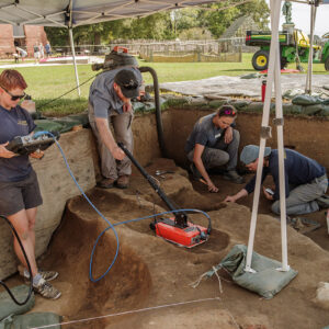 Staff Archaeologist Caitlin Delmas and Director of Archaeology Sean Romo conduct a GPR survey of one of the burials in the 1607 Burial Ground. Senior Staff Archaeologist Anna Shackelford and Staff Archaeologist Natalie Reid conduct excavations at the rear. Natalie's dust pan contains a partial wine bottle she discovered during her digging.