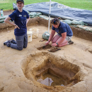 Archaeological Intern Katie Griffith and Archaeological Field Technician Hannah Griffith bisect a posthole near the west-most subfloor pit in the north field. Note the groundwater welling up from below.