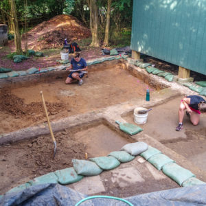 an archaeologist records features while another trowels in a large unit next to a wooden structure