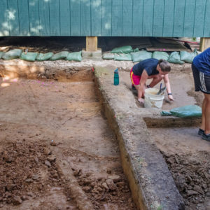 Three archaeologists excavating and documenting features next to a wooden structure