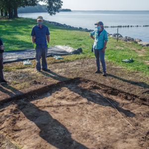 three archaeologists standing on the edges of an excavation unit next to a riverbank