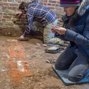 Archaeologist kneels and examines a piece of mortar while another archaeologist excavates in the background