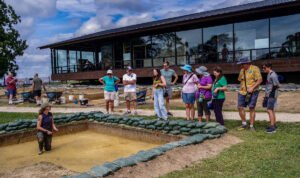 Archaeological Field Technician Ren Willis explains the upcoming burial excavation south of the Archaearium to some visitors. The burial is the dark rectangle just below her in the photo.