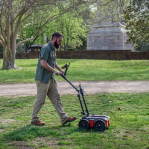man pushing a ground-penetrating radar unit along a transect line in a grassy area
