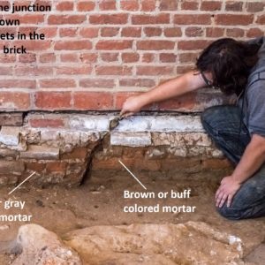 Man points with trowel to mortar between brick wall and excavated dirt floor