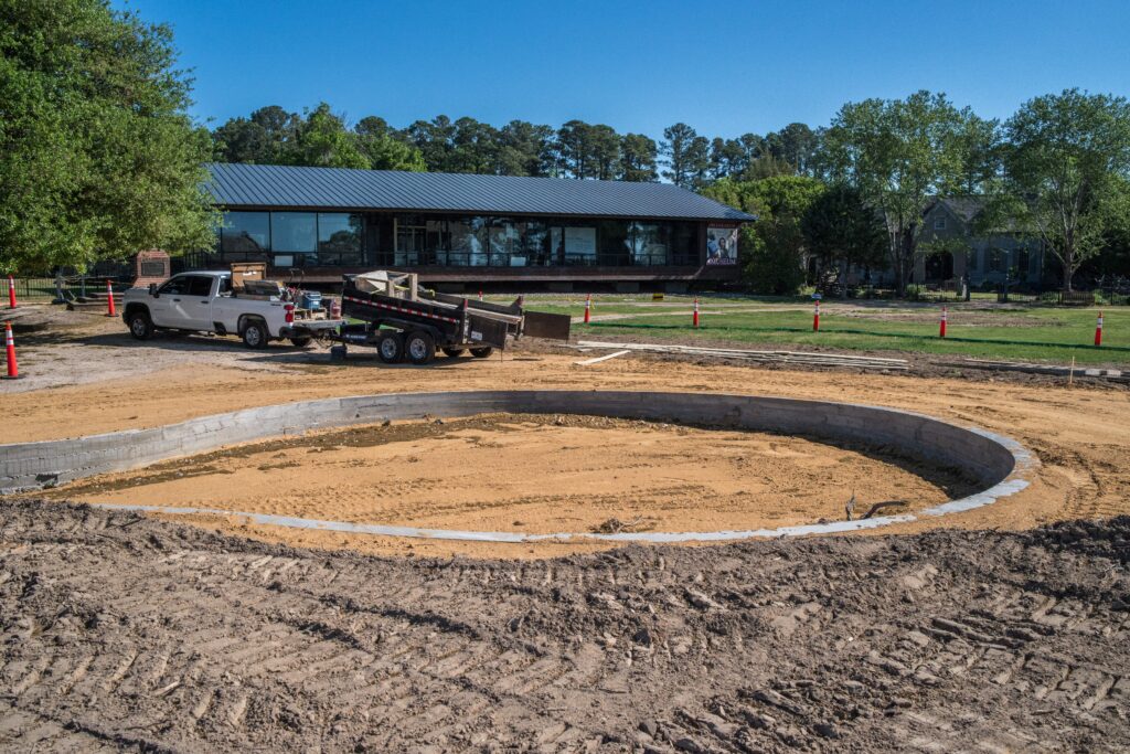 A photo of construction work, with a large, circular space lined with concrete in the foreground, and work trucks in the background near the Archaearium Museum, a single-story glass building.