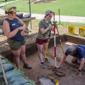 Archaeological Field Technicians Hannah Barch and Josh Barber instruct field school student Grace Blondin-Kissel in the surveying process so as to precisely record the location of archaeological features.