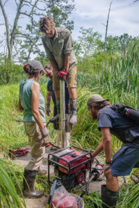 Field school student Connor DeWall puts all his weight on an aluminum tube in an effort to force it down into the swamp as part of the vibracoring process. Field school student Kristin Grossi, TerraSearch Geophysical's Dr. David Leslie, and Archaeological Field Technician Josh Barber assist with the process.
