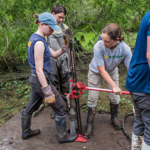 Julia Womersley-Jackman uses a jack to pull a soil sample tube back out of the swamp as part of the vibracoring process. Staff Archaeologist Natalie Reid and field school student Lexy Marcuson assist.