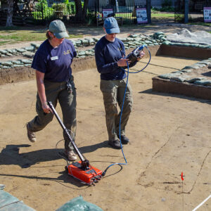 Archaeological Field Technician Eleanor Robb and Staff Archaeologist Natalie Reid perform a GPR survey of the burial excavation area south of the Archaearium. The burials are the long rectangles in the soil (scored in this photo).