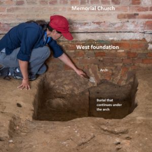Kneeling archaeologist points with trowel to excavated pit in front of brick wall