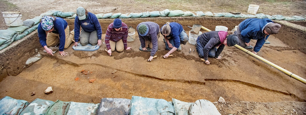 The archaeological team gently scrapes away the top layer of soil in the 1608 palisade extension squares to make features pop for record photography.