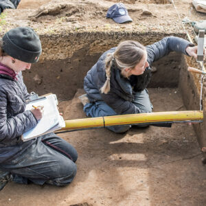 Staff Archaeologist Ren Willis and Archaeological Field Technician Katie Griffith record measurements of different soil layers in the North Church excavations. The yellow conduit houses telecommunications cable.