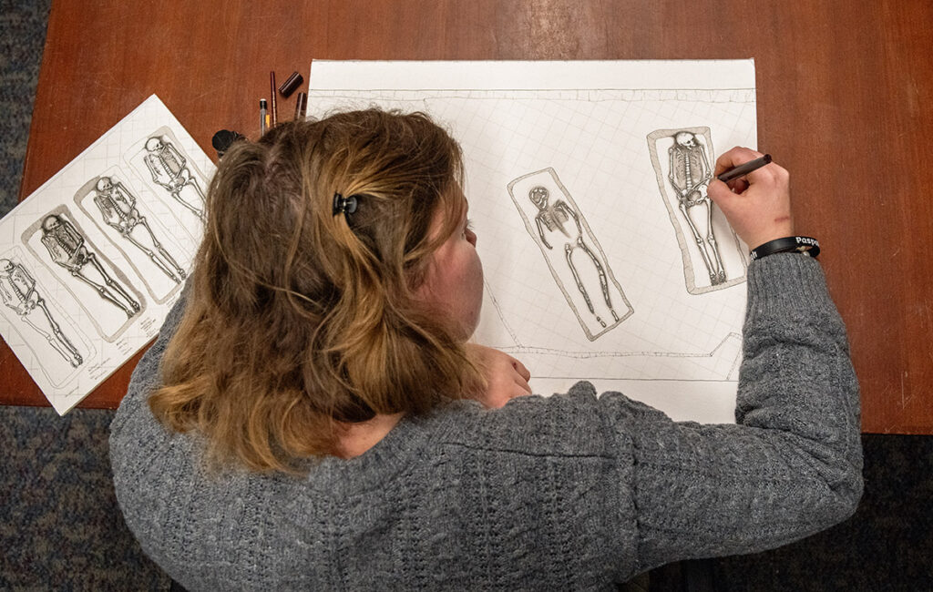 Archaeological Field Technician Eleanor Robb draws the human remains found during 2024's excavations at the 1607 burial ground. At left is a sampler showing different styles of representation that she presented to the archaeological team.