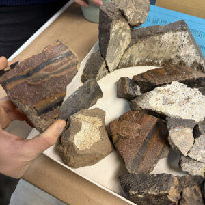 The newly discovered sherd of melting pot is held and compared to an existing partial jar already in the collection. The new piece likely belongs to the same vessel.
