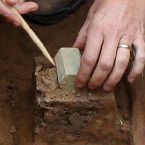 tarnished, oblong artifact being removed from the soil with a wooden pick