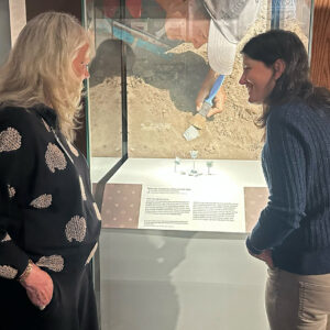Jamestown-Yorktown Foundation Senior Curator Beverly (Bly) Straube and Senior Staff Archaeologist Mary Anna Hartley discuss one of the cases in the "Following the Dragon" exhibit. The case's background photo shows Mary Anna excavating one of the pieces of the wine cup on exhibit.