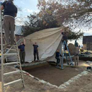 In order to get a record shot with even lighting, the team blocks the sun while Senior Staff Archaeologist Dr. Chuck Durfor climbs a ladder to take the photo.