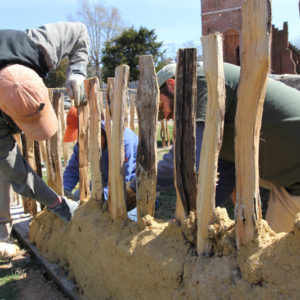 three people applying mud to a row of upright wooden studs