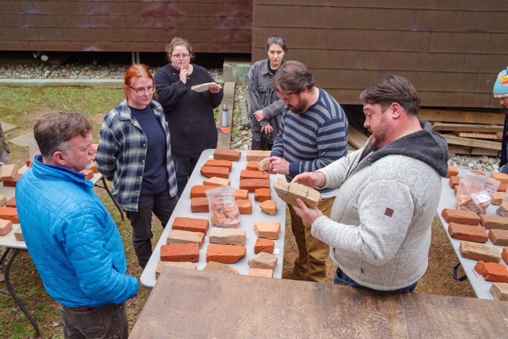 Six people stand around a table on which lie several bricks. The people are examining the bricks.