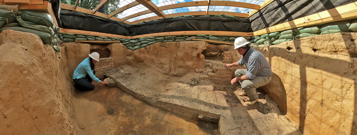 two archaeologists kneeling in a large unit covered with a wooden platform
