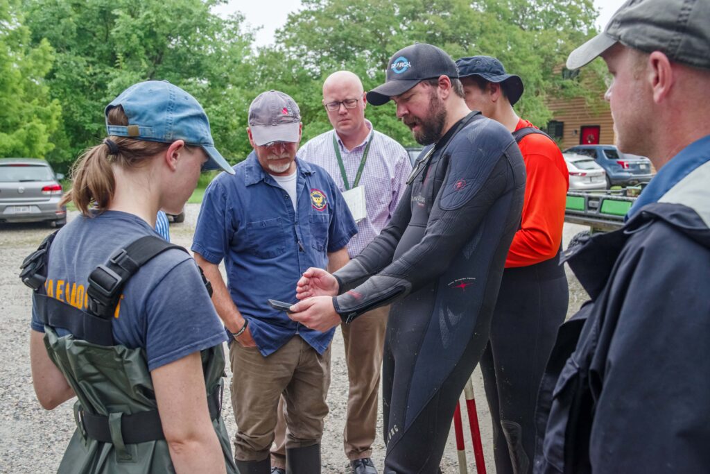 A group of people stand around a man in a wetsuit who is holding a tablet. Everyone is looking at the tablet. 