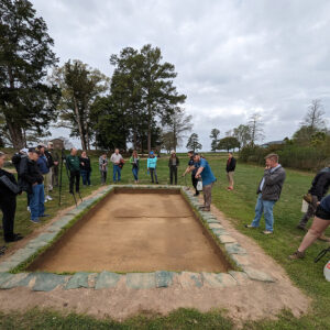 Senior Staff Archaeologist Sean Romo explains the features in the north field excavations. The pit feature is the round stain at left center. The ditch feature is the dark stain running from top to bottom (west to east) near the right edge of the feature.