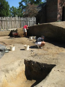 Excavated ditch feature in the foreground with two archaeologists troweling and shoveling in the background