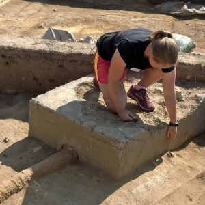 archaeologist kneeling on an excavated septic tank and pointing downwards at an in situ ceramic sherd