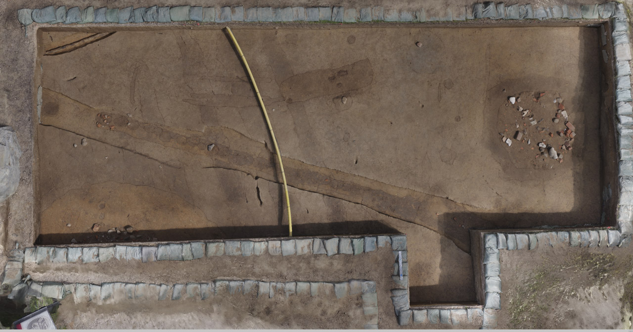 An overview of the North Church excavations. The 1608 palisade extension runs left to right through the photo, with individual postholes scored by the archaeologists. The well is the circular feature at the right of the photo, filled with bricks, limestone, and coral. The possible other well is at the bottom left of the photo, only partially revealed, also containing architectural materials. A burial is visible at the top center of the photo. The yellow pipe is conduit for telecommunications wire. Faint darker lines, roughly paralleling the conduit, are planting furrows.