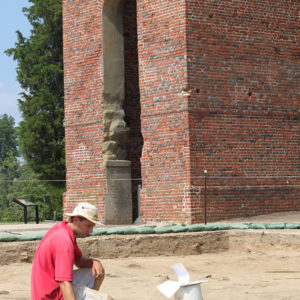Archaeologist kneeling in an excavation area in front of a brick church tower