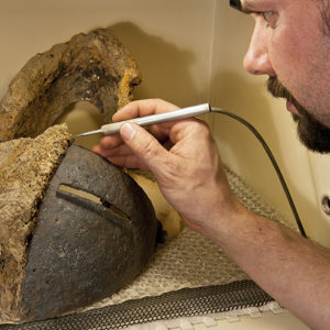 conservator using an electric tool to clean a half-conserved iron helmet