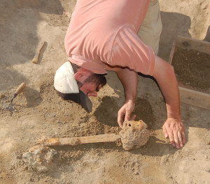 An archaeologist bends over to examine a broadsword lying in situ