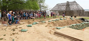 archaeologist giving a tour to a large group of visitors in front of an excavation area and a wooden building frame