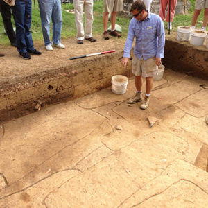 archaeologist standing in an excavation area and pointing out outlined features to a gathered group