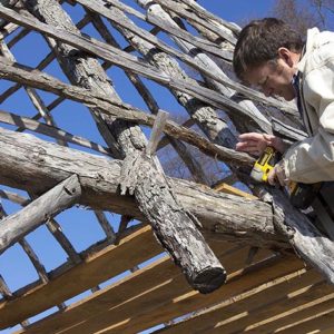 man using a drill to secure horizontal planks into the roof of a wooden structure