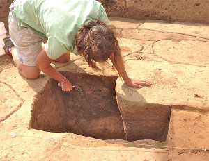 archaeologist bending over an excavation unit and scraping the sides with a trowel