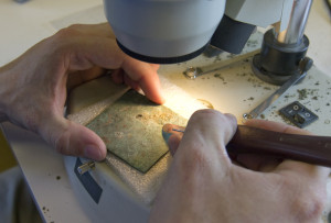 Scalpel being used to clean a small, square piece of copper with a small hole in the top center