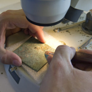 Scalpel being used to clean a small, square piece of copper with a small hole in the top center
