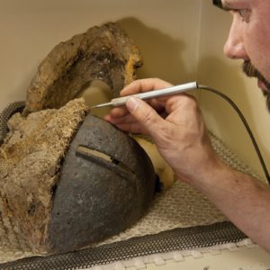 Conservator using a pick to remove corrosion from a half-conserved helmet