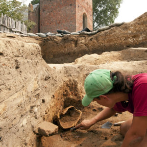 archaeologist using a trowel to scrape soil from around an in situ corroded helmet