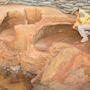 archaeologist kneeling in an excavation area lined with sandbags and containing two brick oven features