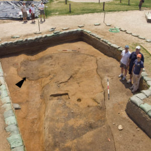 three archaeologists standing in an excavation unit with a feature marked by an overlaid arrow