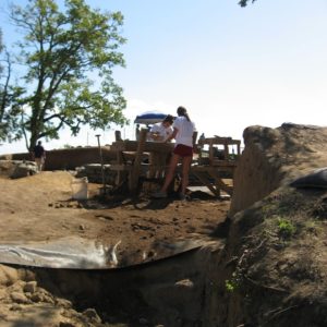 Partially-uncovered tarp within an excavation unit with view of two people screening artifacts in the background