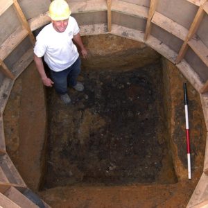 Archaeologist standing in excavated square-shaped well surrounded by circular supporting wooden frame